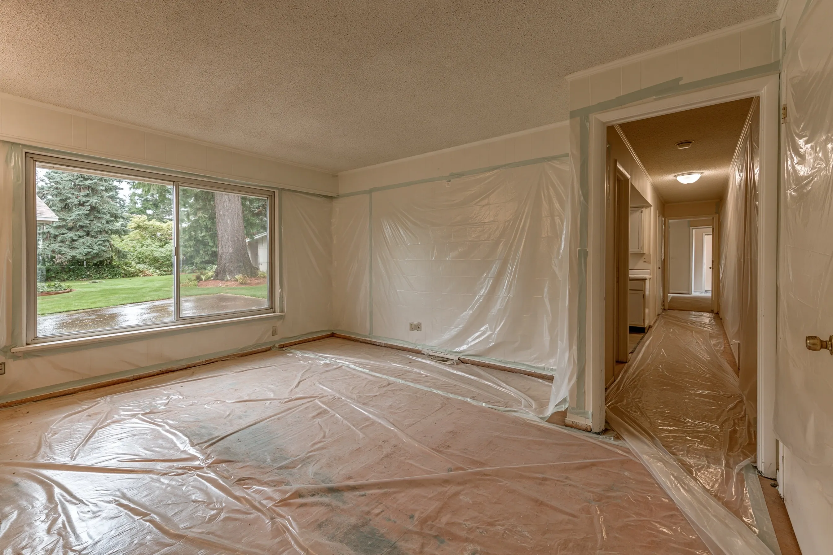 Empty 1970s rambler with popcorn ceilings through the living room and hall, bare floors and plastic sheeting rolled out