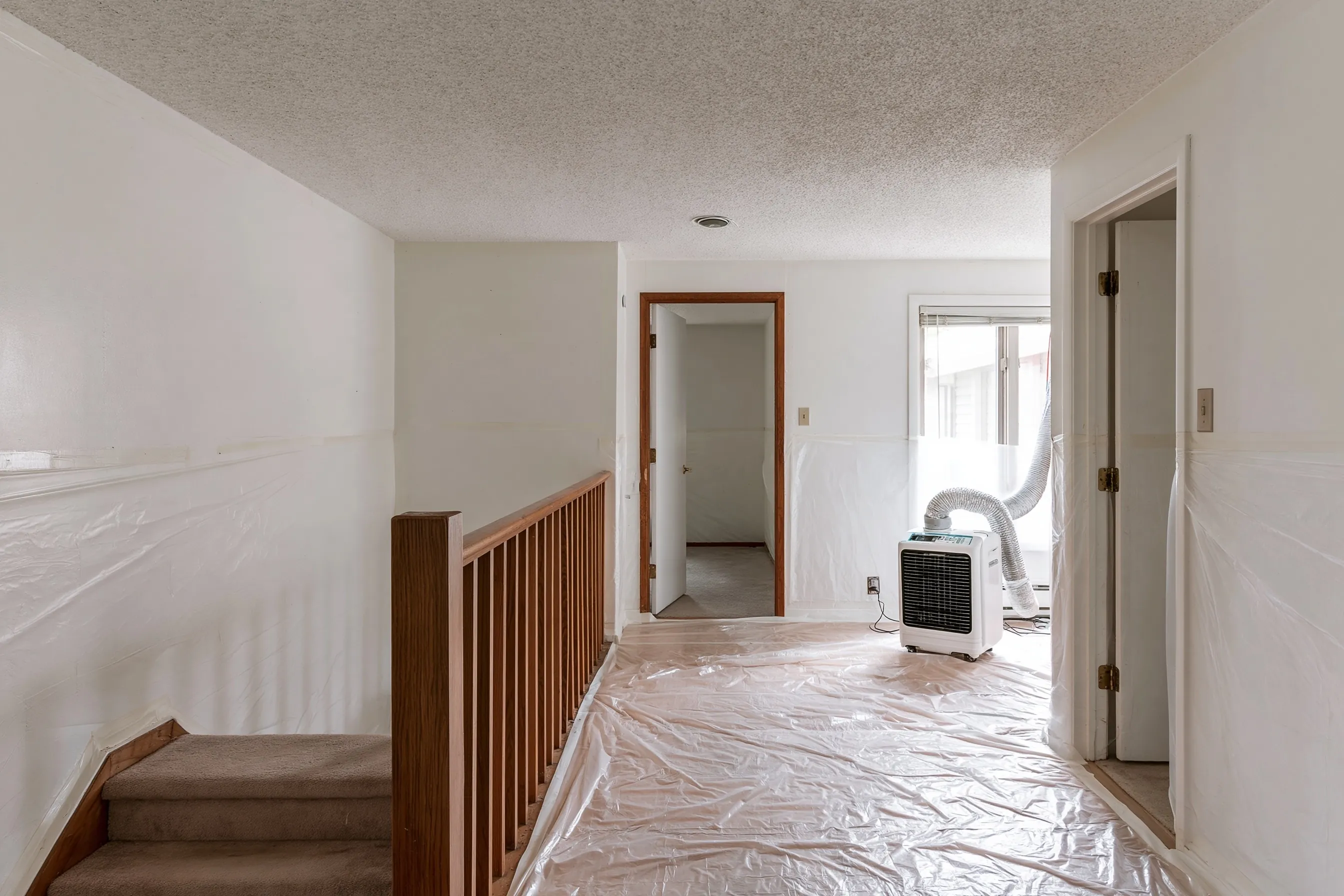 Upper floor of a 1960s split-level with a painted popcorn ceiling, plastic over the floor and walls, and the half-flight stair